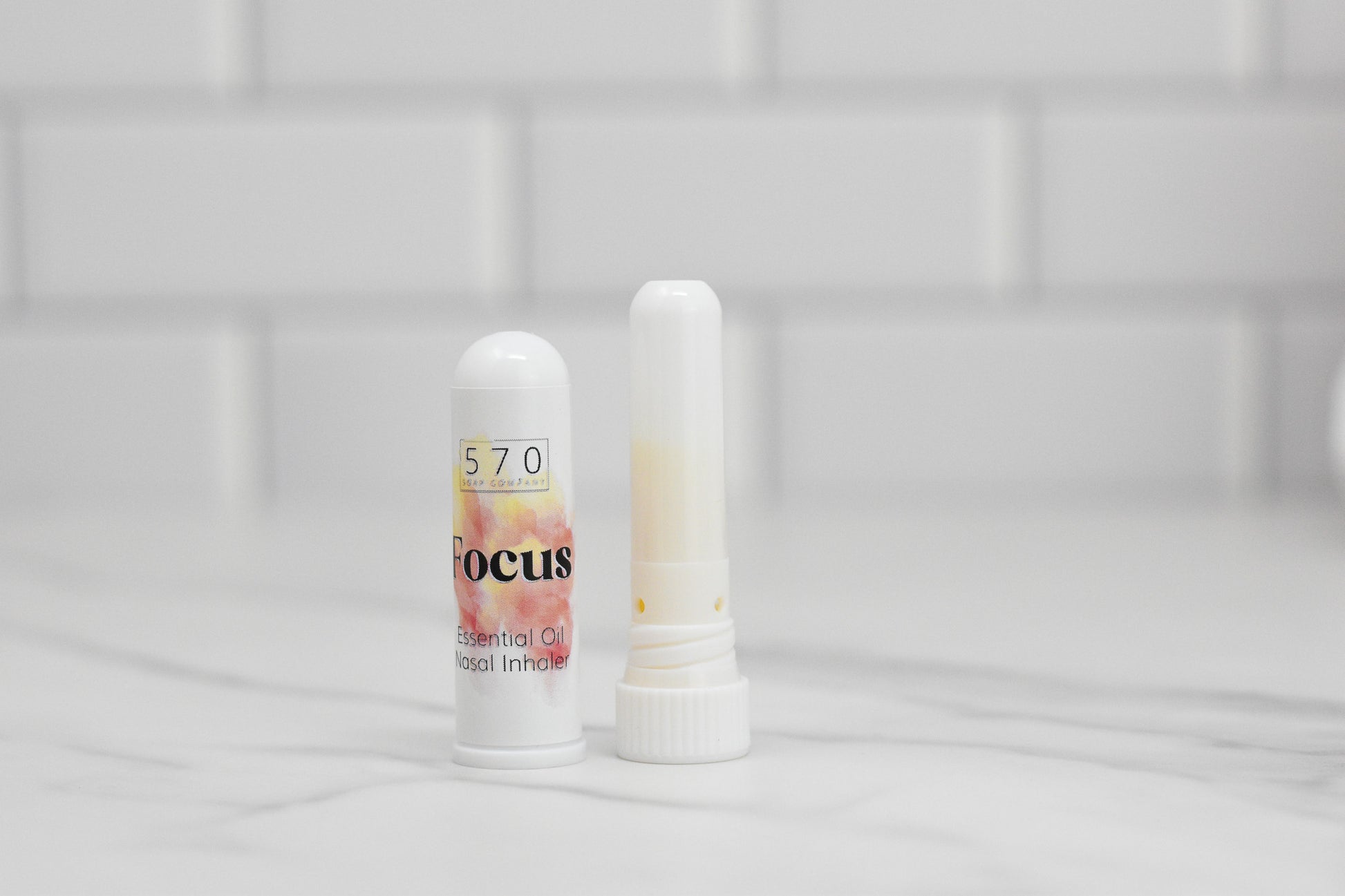 A white Nasal Inhaler from 570 Soap Co., labeled Focus, sits on a marble surface with its cap removed to reveal the tip. Positioned against a white tiled background, this natural relief tool is crafted for those times when you need concentration and clarity.