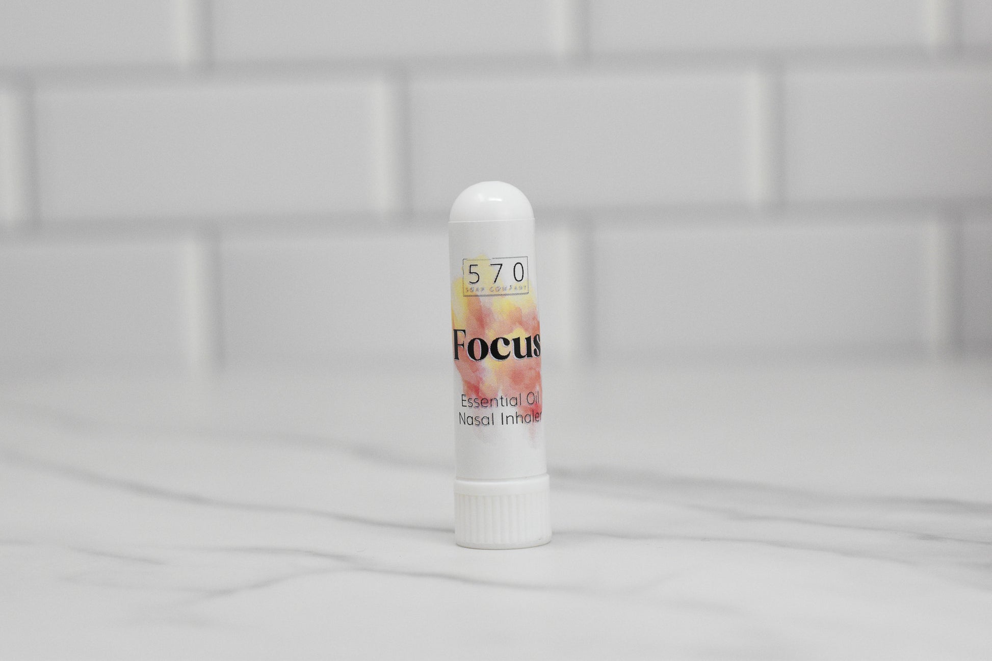 A small white inhaler labeled 570 Soap Co. Nasal Inhaler stands on a marble surface, offering natural relief. The label includes "Focus," "Essential Oil Nasal Inhaler," and features a floral design. The background of white subway tiles highlights its blend of essential oils for soothing comfort.