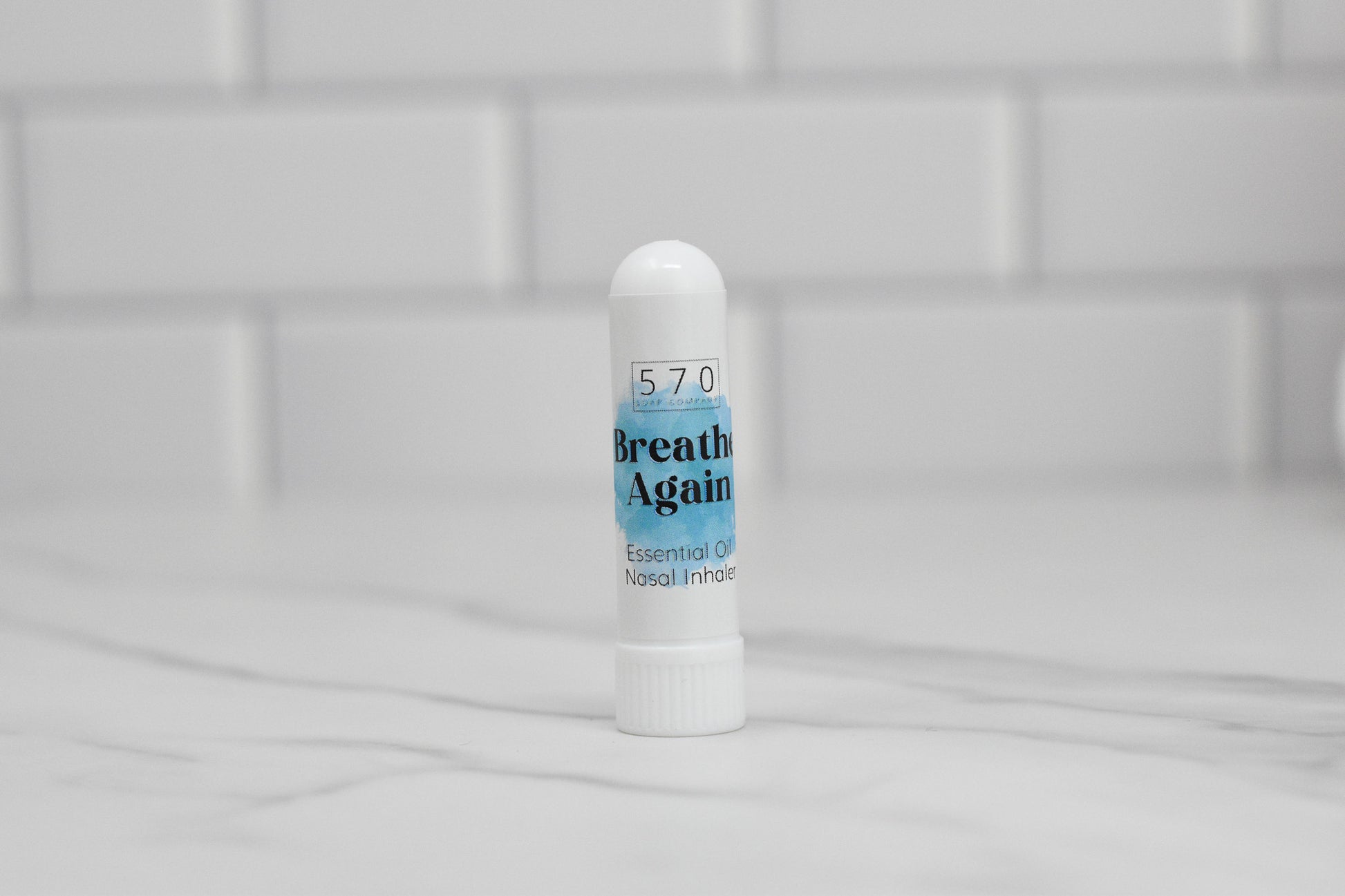 A white cylindrical nasal inhaler labeled 570 Soap Co. Nasal Inhaler stands on a marble countertop with a white tiled backsplash, offering natural relief through the power of essential oils.