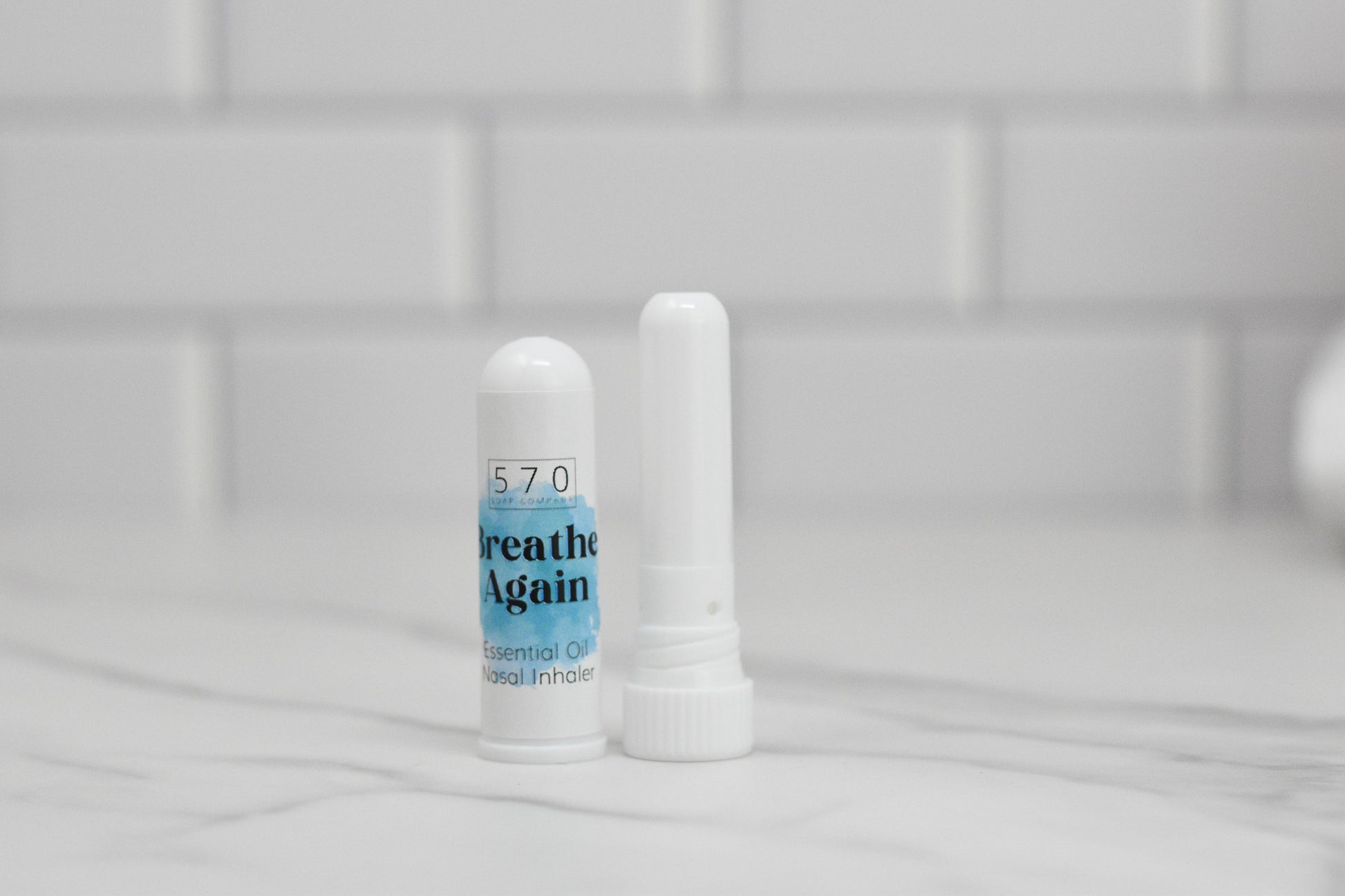 Two white cylindrical Nasal Inhalers from 570 Soap Co., infused with essential oils, rest on a white tiled surface. One carries the label Breathe Again. A sleek backdrop of white tiles enhances the minimalist setting, promising natural relief in every breath.