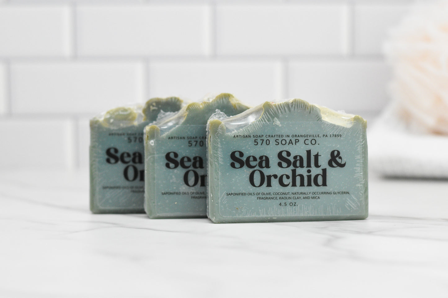 Three luxurious bars of Sea Salt & Orchid Bar Soap: Coastal Tranquility from 570 Soap Co. rest on a white marble surface against a white tiled background. The soap, with its greenish hue and textured top edge, promises an indulgent and refreshing lather.