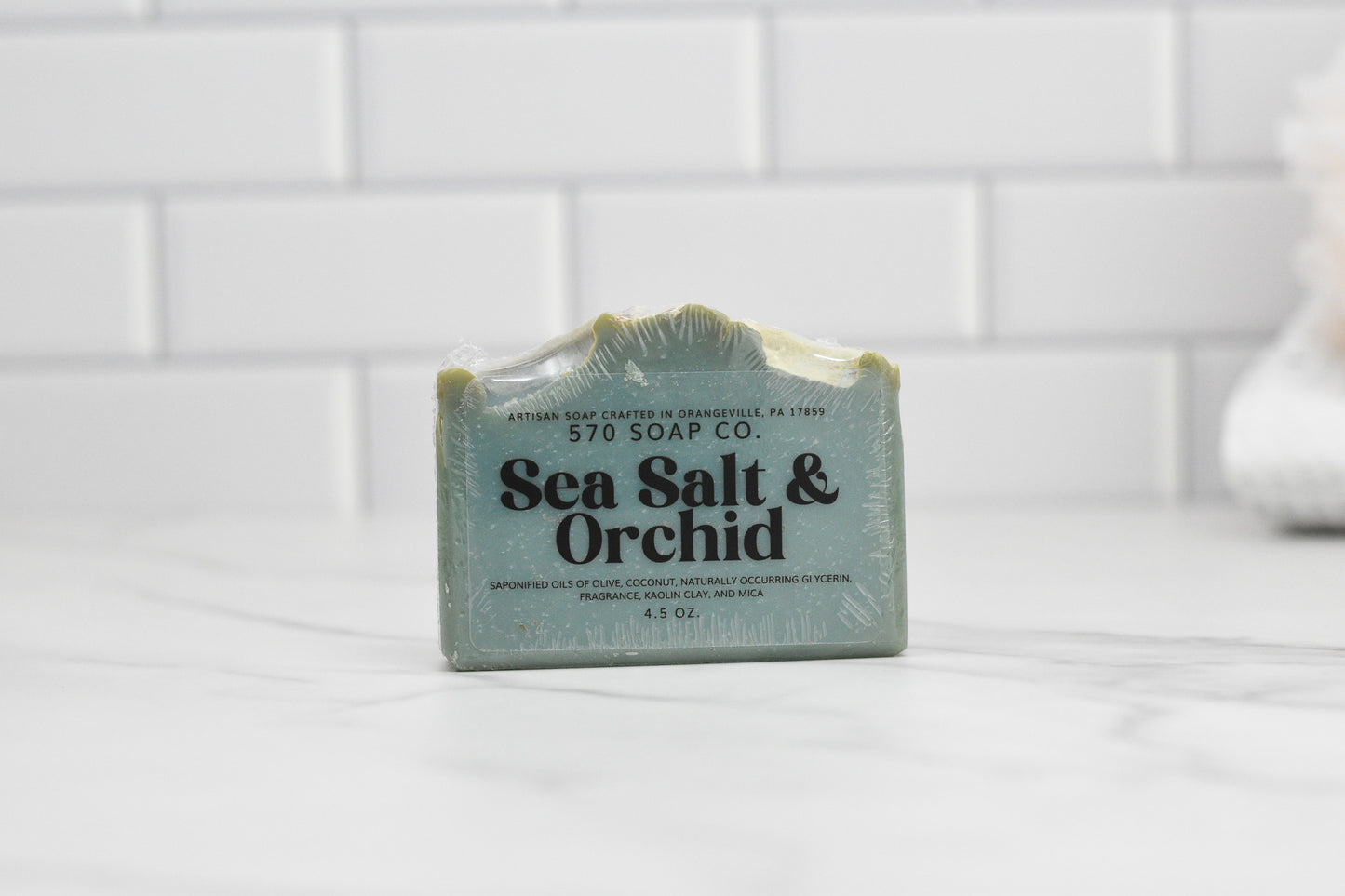A bar of Sea Salt & Orchid Bar Soap: Coastal Tranquility from 570 Soap Co. is elegantly displayed on a white marble surface against a white tiled wall. The packaging, with its simple design, unfolds the promise of a luxurious lather, featuring product details gracefully printed on it.