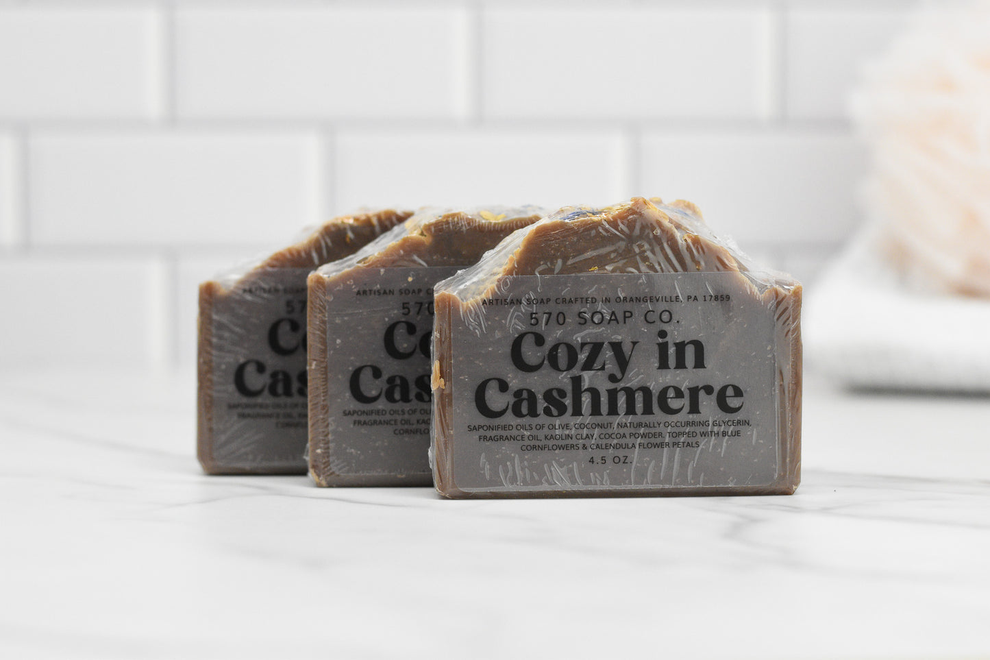Three bars of 570 Soap Co.'s Cozy in Cashmere Bar Soap: Luxurious Blend, infused with nurturing oils, are set on a white marble countertop against a white tiled wall. The soap bars are wrapped in plastic, and the text on the label is clearly visible.