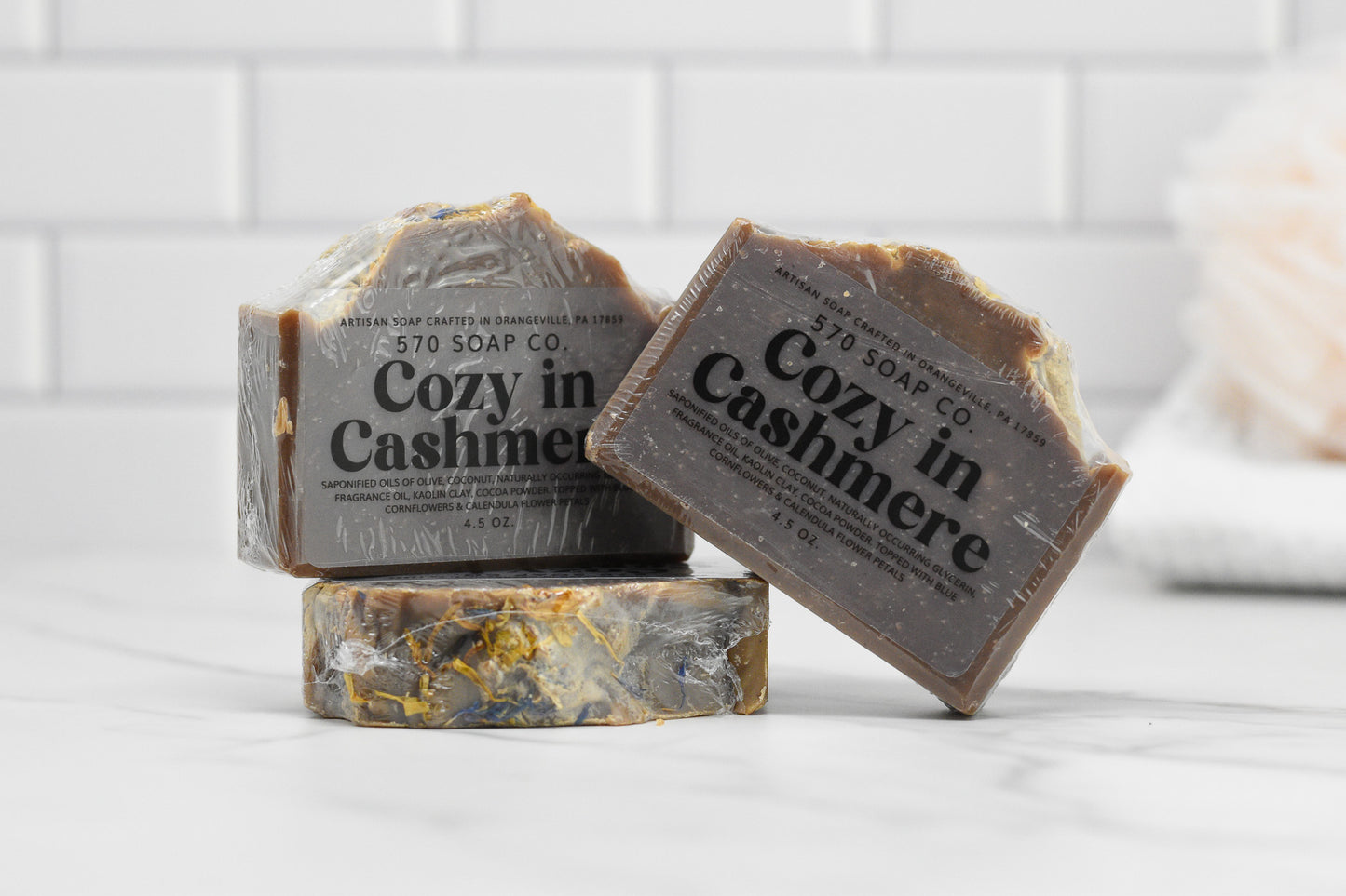 Two bars of Cozy in Cashmere Bar Soap: Luxurious Blend by 570 Soap Co. are displayed against a tiled background. One bar stands upright on two others that are stacked, showcasing its marbled texture with dark and light swirls that create a scent symphony, enriched by nurturing oils for a luxurious feel.