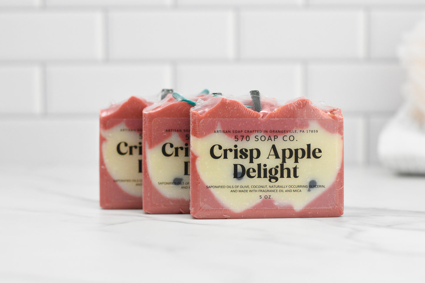 Displayed on a white marble surface against a white tiled backdrop are three bars of 570 Soap Co.'s Crisp Apple Delight Bar Soap. Enriched with nourishing oils, each bar is individually wrapped in a red and cream color scheme that enhances its natural appeal.