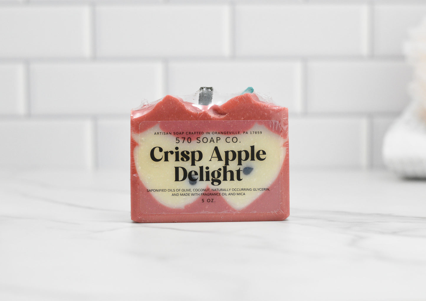 A bar of Crisp Apple Delight Bar Soap from 570 Soap Co., weighing 5 oz, is infused with nourishing oils. The red soap boasts a white center and rests against a white tiled background.