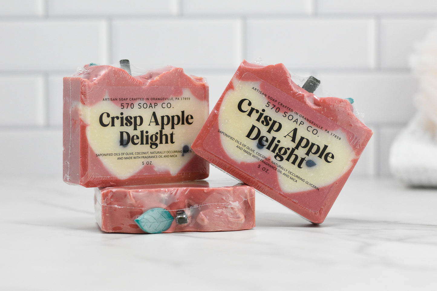 Three bars of 570 Soap Co.'s Crisp Apple Delight Bar Soap are stacked on a white surface. The pink soap, enriched with nourishing oils, features a textured top that mimics apple skin, each piece topped with a small decorative apple stem. A backdrop of white tiles enhances the apple-inspired fragrance visual.