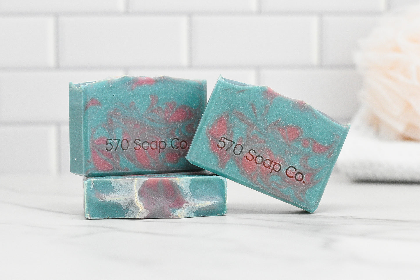 Three bars of the Peony & Amberwood Bar Soap from 570 Soap Co., featuring teal and pink marbling and made with nourishing ingredients, are arranged on a white surface against a tiled background. The captivating fragrance fills the air, while a white loofah is visible in the backdrop.