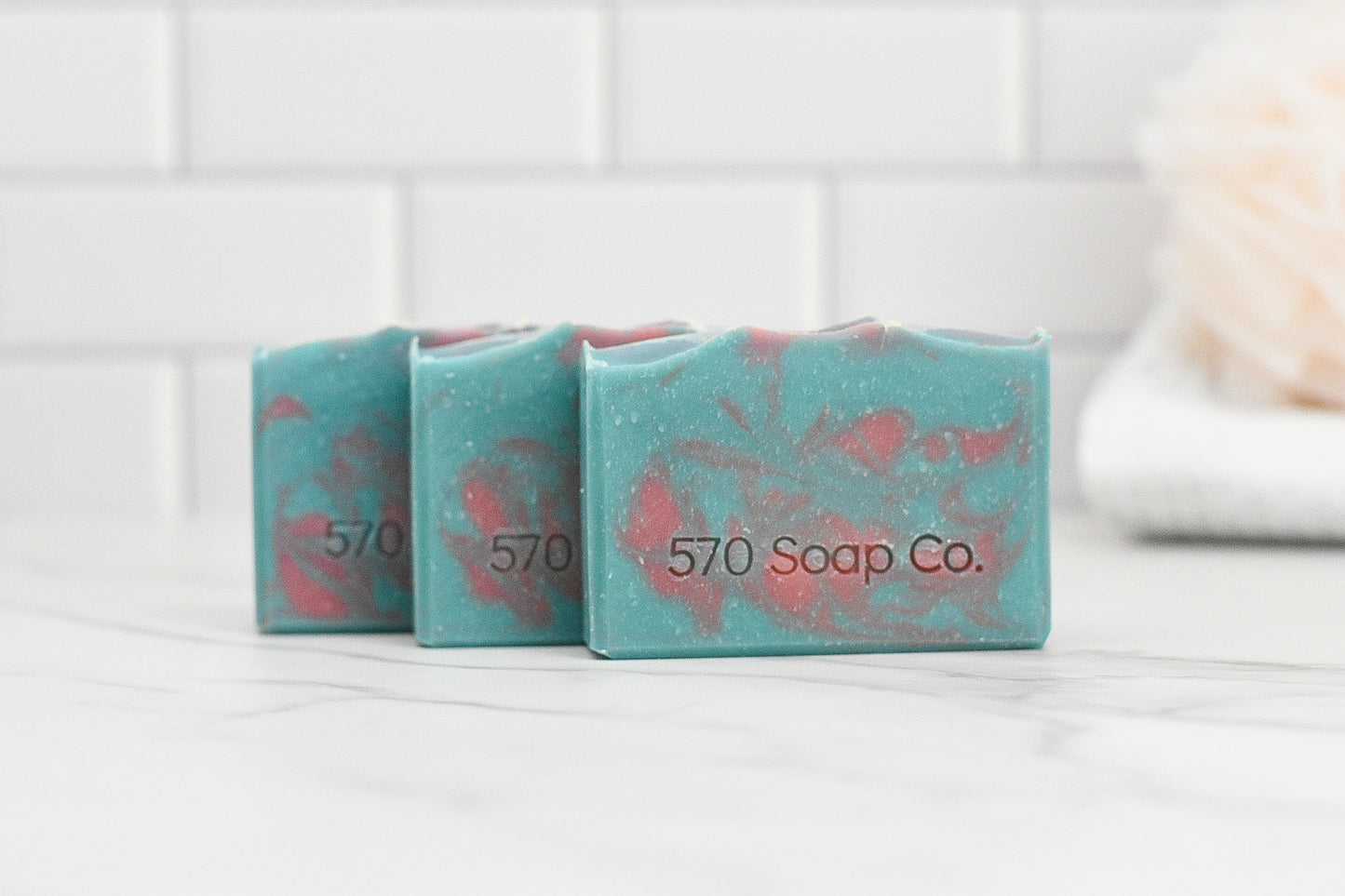 Three bars of Peony & Amberwood Bar Soap by 570 Soap Co. are arranged on a marble surface, highlighting their striking blue and pink marbled design. Formulated with nurturing ingredients, these soaps offer an enchanting aroma, as a gently blurred bath sponge lies nearby against the backdrop of a white tile wall.