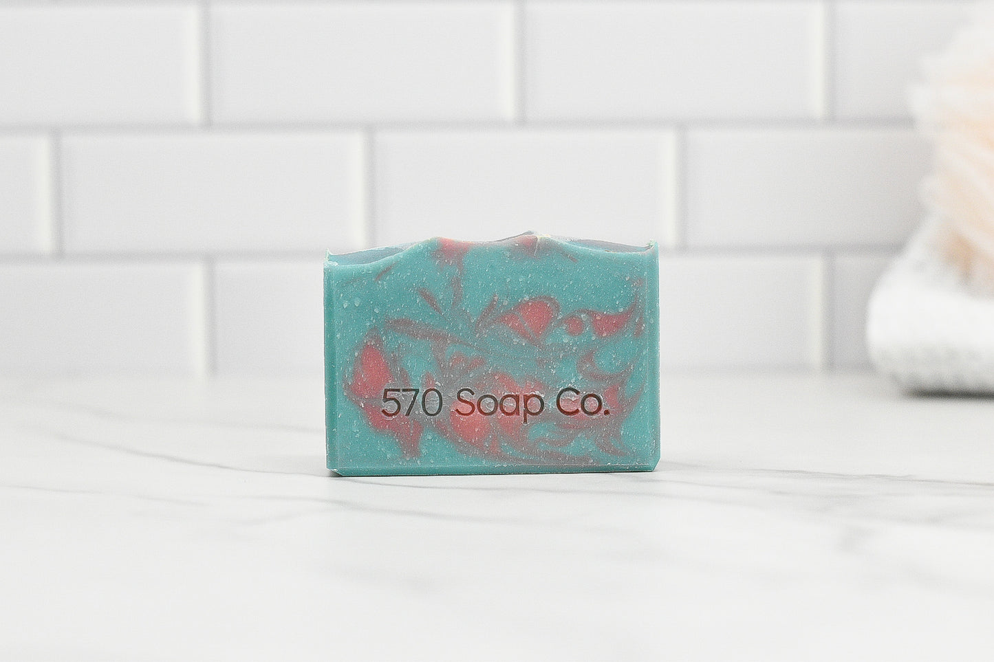 A Peony & Amberwood Bar Soap by 570 Soap Co., featuring a vibrant turquoise and pink marbled design, rests on a white marble surface. Surrounded by white subway tiles and a softly blurred towel in the background, this soap bar offers nourishing ingredients and an enchanting fragrance.