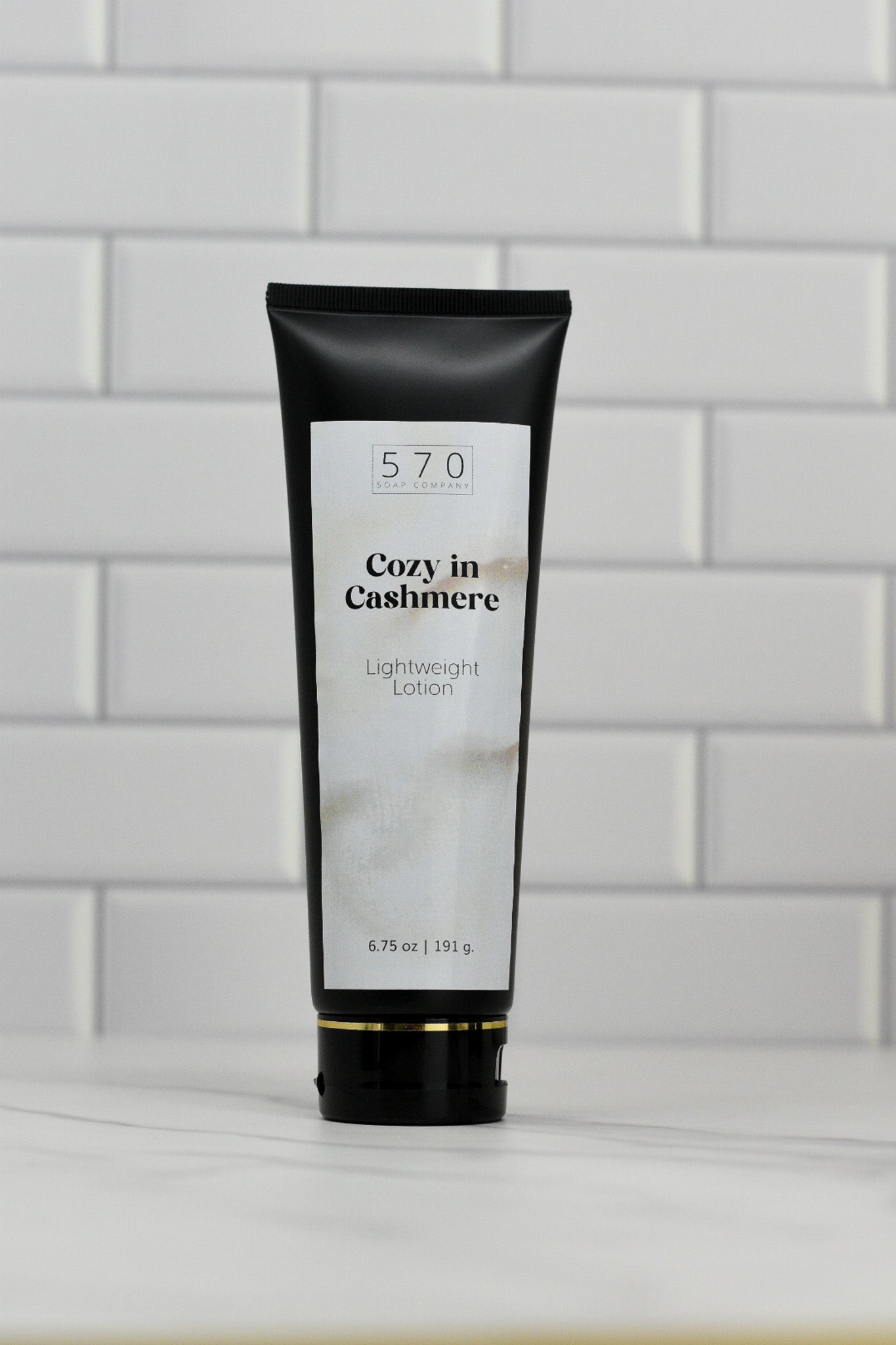 A sleek black tube with a white label featuring the 570 Soap Co.'s Cozy in Cashmere Lightweight Lotion stands upright on a surface against a white tiled wall. Weighing in at 6.75 oz (191.9 g), this lotion offers an enchanting fragrance and nourishing ingredients, promising a luxurious touch to your skincare routine.