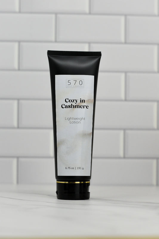 A sleek black tube with a white label featuring the 570 Soap Co.'s Cozy in Cashmere Lightweight Lotion stands upright on a surface against a white tiled wall. Weighing in at 6.75 oz (191.9 g), this lotion offers an enchanting fragrance and nourishing ingredients, promising a luxurious touch to your skincare routine.