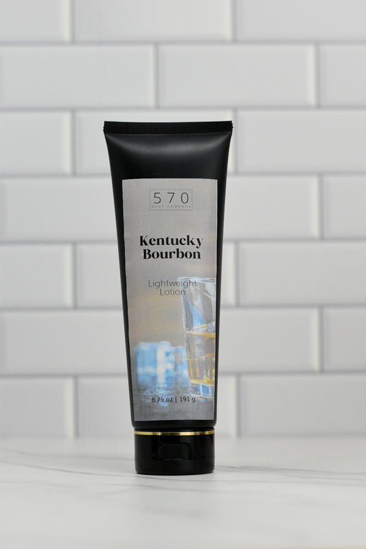 A black tube of 570 Soap Co.'s Kentucky Bourbon Lightweight Lotion stands upright against a white tiled background, exuding a sophisticated fragrance. The label showcases an image of a glass of bourbon, hinting at its nourishing ingredients. The tube holds 6.5 oz (191 g) of lotion.