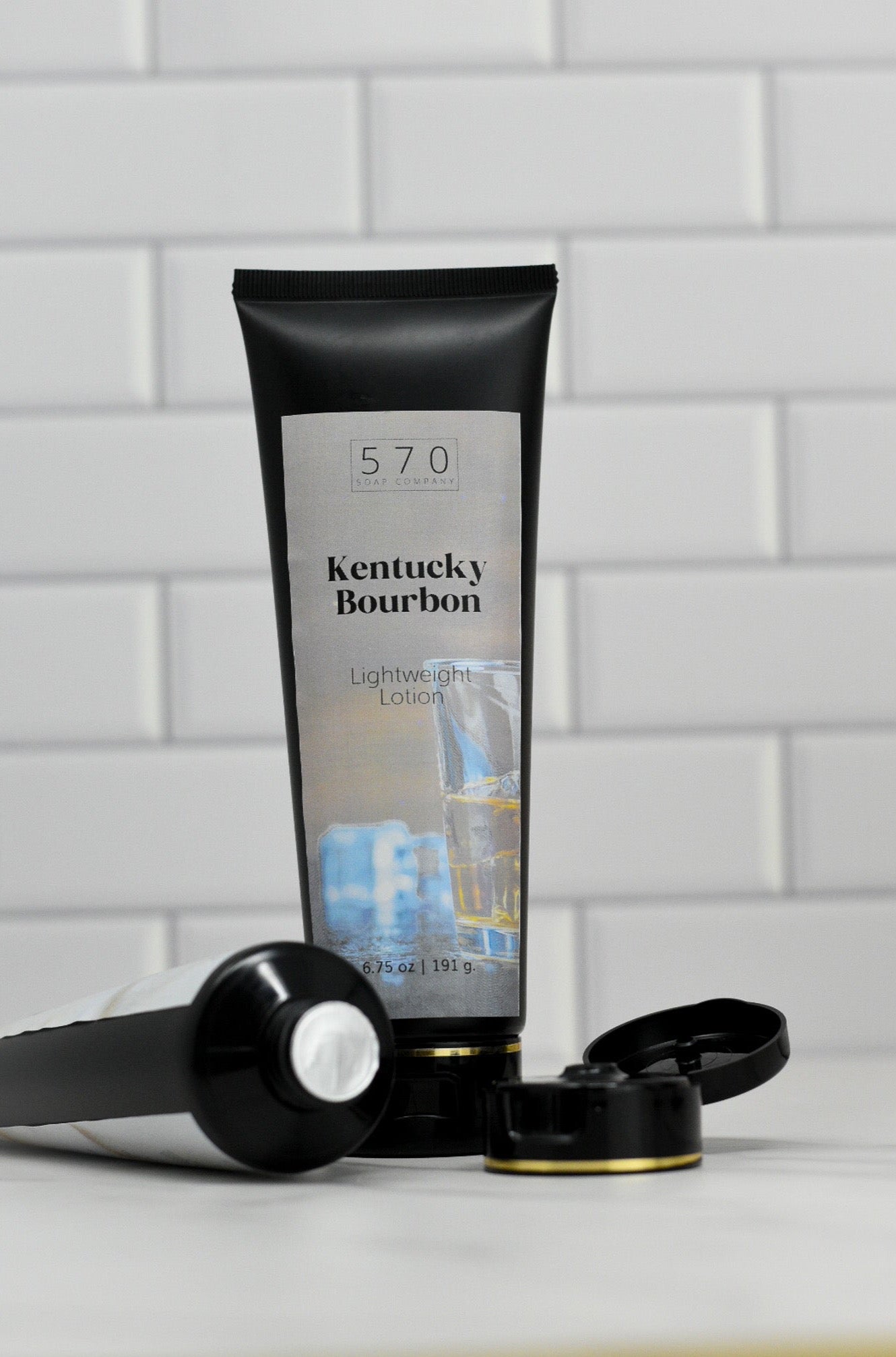 Two tubes of Kentucky Bourbon Lightweight Lotion by 570 Soap Co. are placed against a white tiled wall, embodying a sophisticated fragrance. The design includes imagery of ice cubes and a whiskey glass, suggesting the nourishing ingredients found in this lightweight lotion.