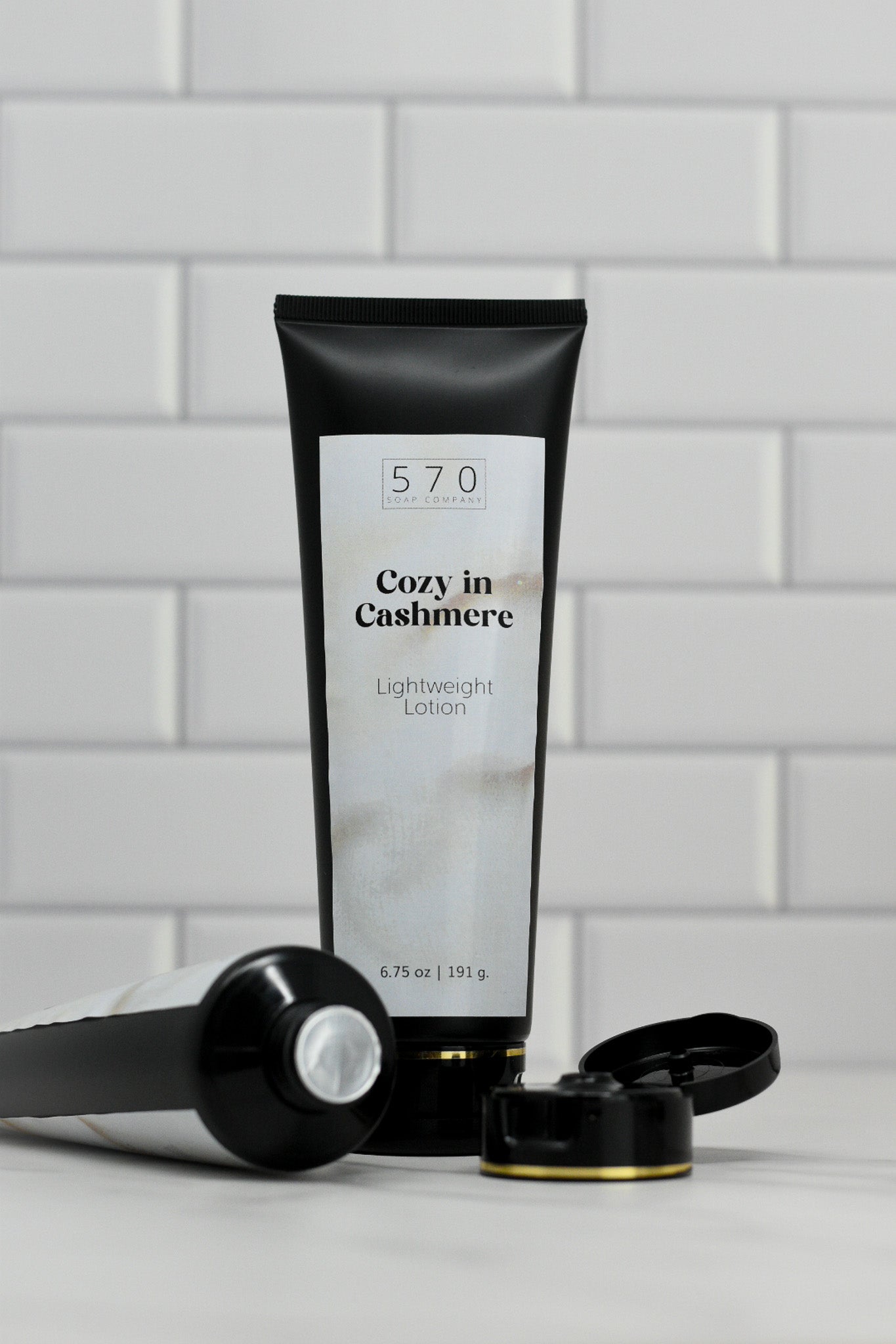 A tube of 570 Soap Co.'s Cozy in Cashmere Lightweight Lotion, featuring a black cap, stands upright against a white tiled background. Nearby, another tube from the same brand, known for its nourishing ingredients and enchanting fragrance, lies horizontally with its cap off.