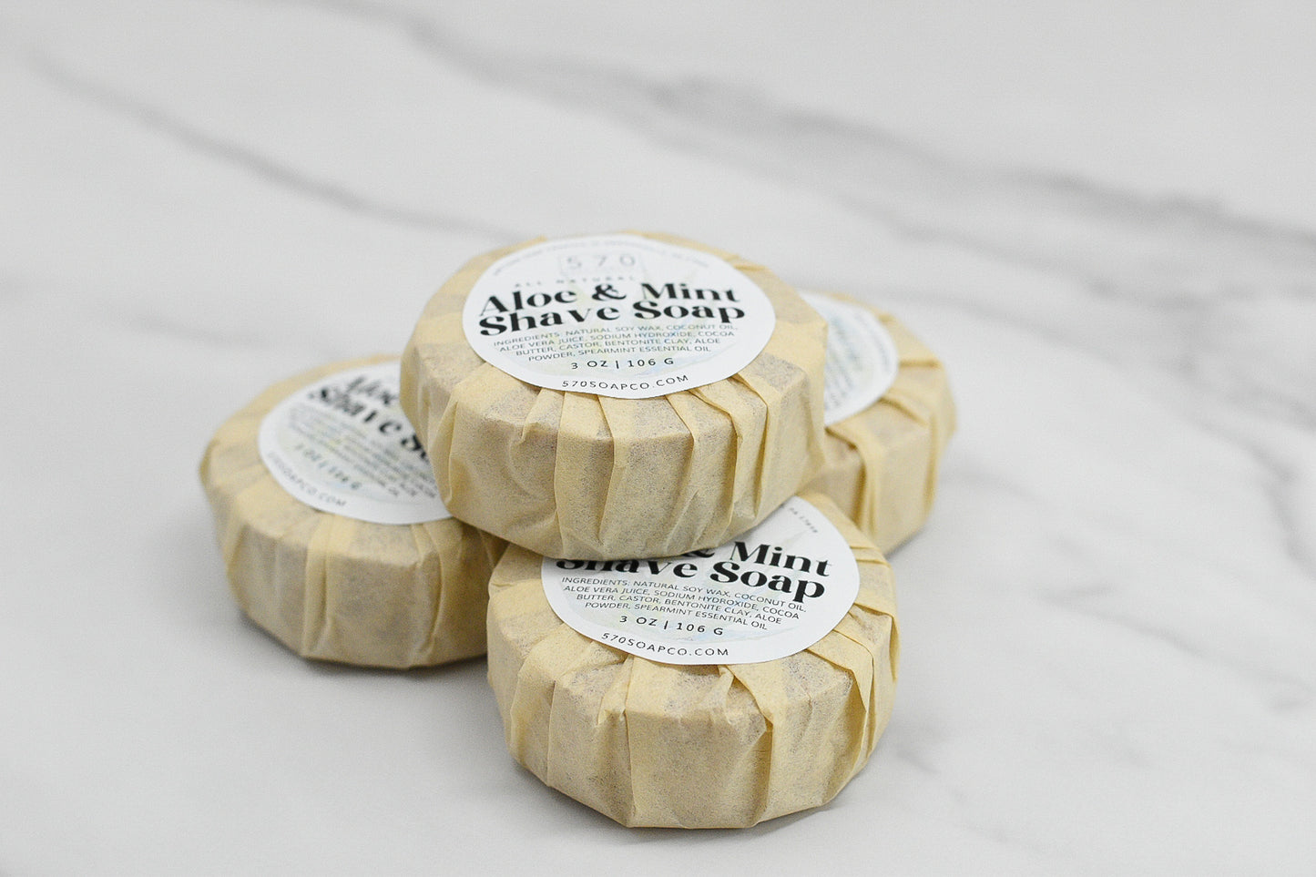 Four wrapped bars of Shave Soap - Luxurious Smoothness for Him & Her by 570 Soap Co. are stacked on a white marble surface. Each circular soap is encased in beige paper with a label displaying the product name, website, and ingredients.