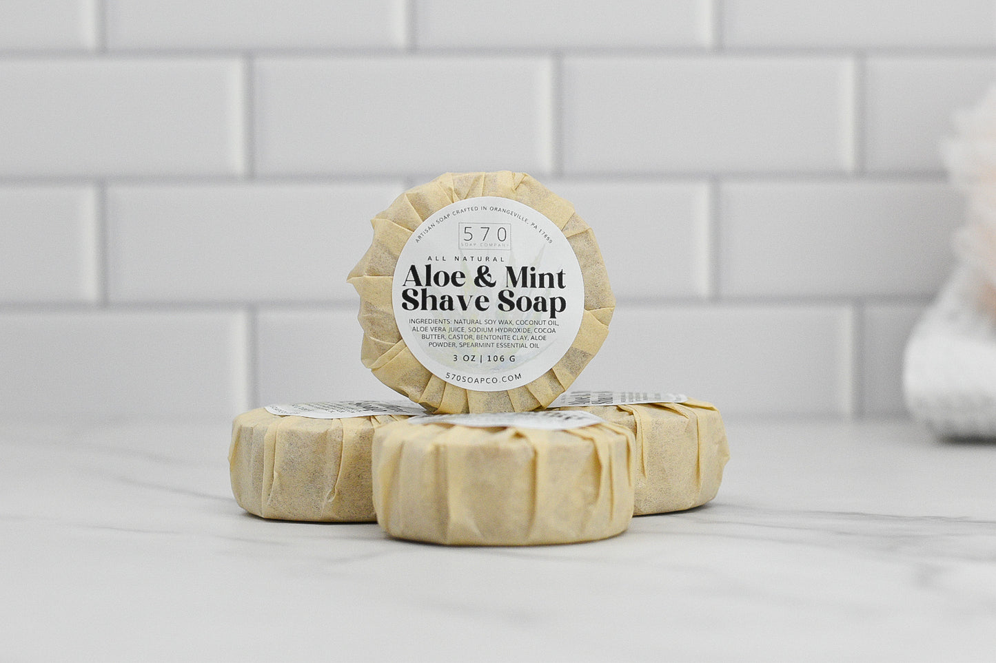Three round bars of Shave Soap - Luxurious Smoothness for Him & Her by 570 Soap Co. are elegantly stacked on a marble surface. Each bar is wrapped in beige paper, showcasing labels that highlight their all-natural ingredients and 3 oz size, set against a backdrop of white subway tiles.
