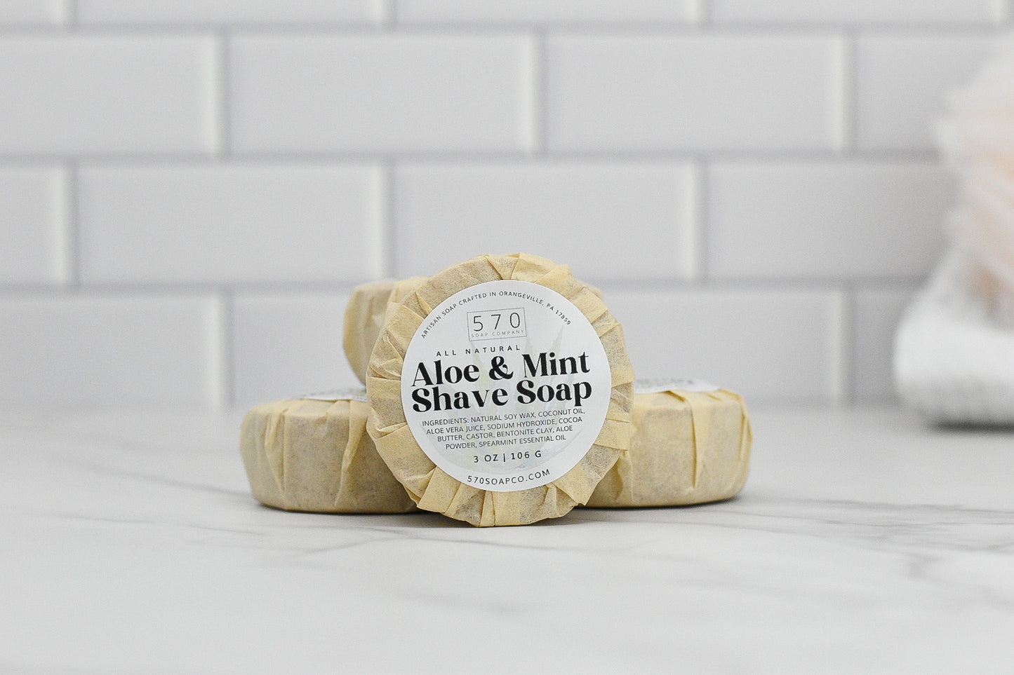Three wrapped bars of Shave Soap - Luxurious Smoothness for Him & Her from 570 Soap Co., featuring a luxurious blend of natural ingredients and soothing properties, are neatly stacked on a marble surface. The backdrop showcases a white tiled wall, creating a clean and minimalist aesthetic.