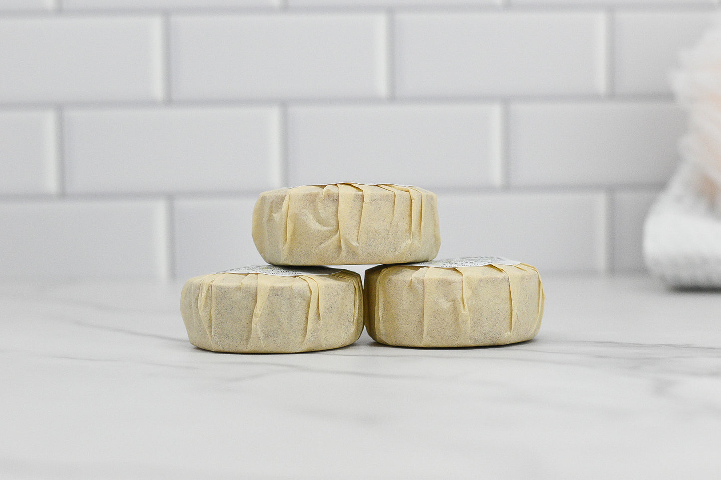 Three round bars of Shave Soap by 570 Soap Co., wrapped and crafted from a luxurious blend of natural ingredients, stacked on a marble surface against a white tile background.