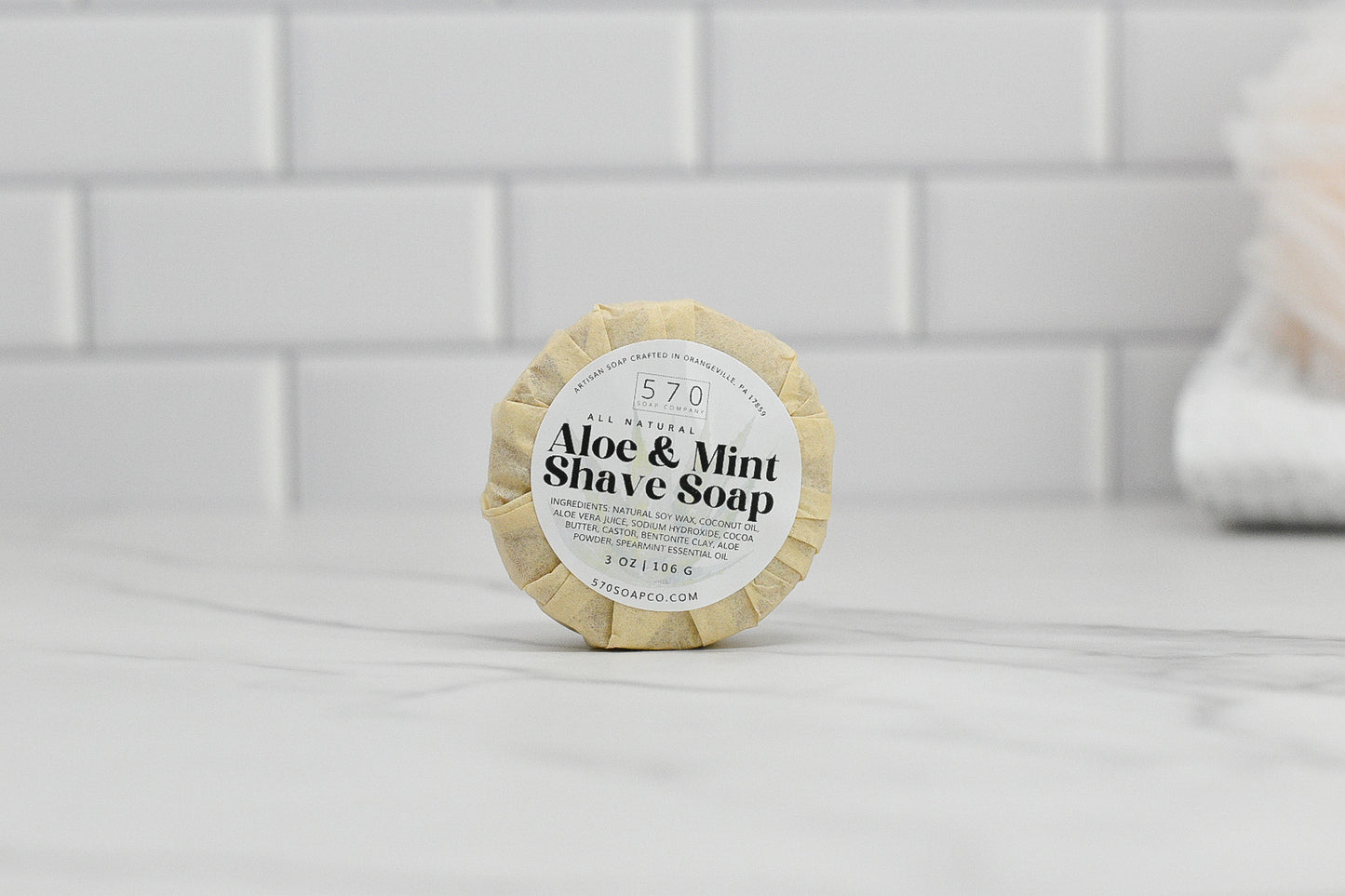 A round bar of 570 Soap Co.'s Shave Soap - Luxurious Smoothness for Him & Her, enveloped in beige paper, rests elegantly on a white marble surface. The scene is complemented by a white tiled wall in the background and a blurred white towel, accentuating its natural ingredients.
