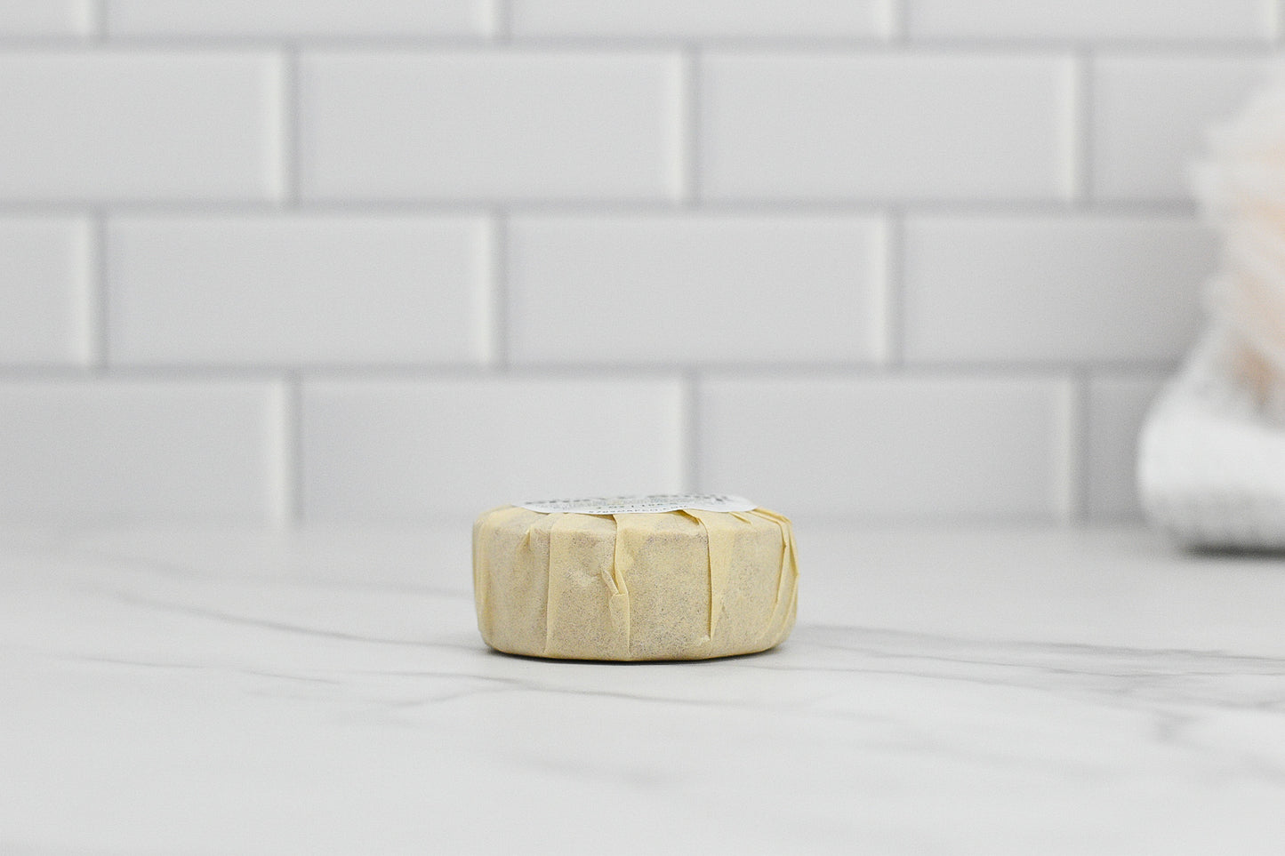 A wrapped Shave Soap - Luxurious Smoothness for Him & Her by 570 Soap Co., featuring natural ingredients, sits on a marble countertop against a white subway tile backsplash. A blurred towel in the background enhances the clean and minimalist bathroom ambiance.