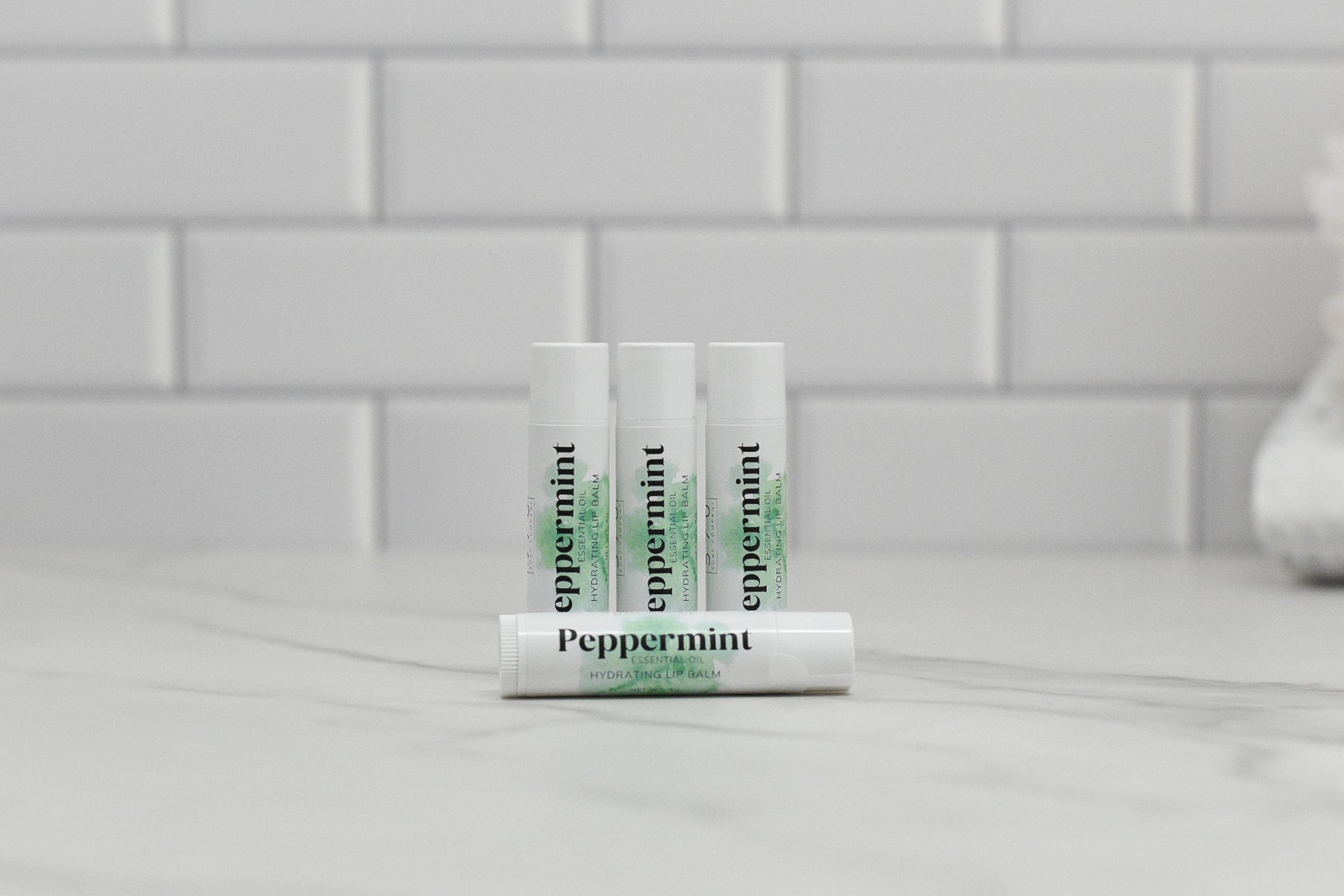 Four tubes of 570 Soap Co.'s Peppermint Essential Oil Lip Balm, all in white, are arranged on a marble countertop against a backdrop of white subway tiles. Three tubes are positioned upright while one rests horizontally in front, offering hydration and nourishment for your lips.