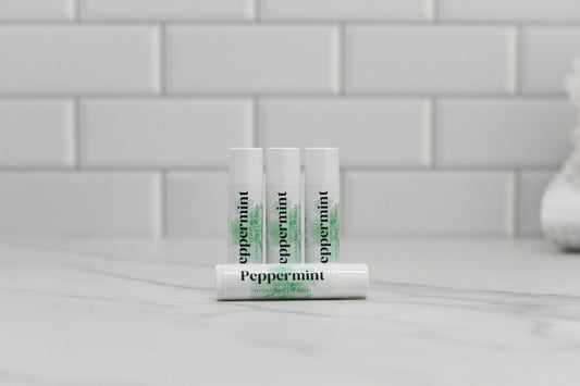 Four tubes of 570 Soap Co.'s Peppermint Essential Oil Lip Balm, all in white, are arranged on a marble countertop against a backdrop of white subway tiles. Three tubes are positioned upright while one rests horizontally in front, offering hydration and nourishment for your lips.