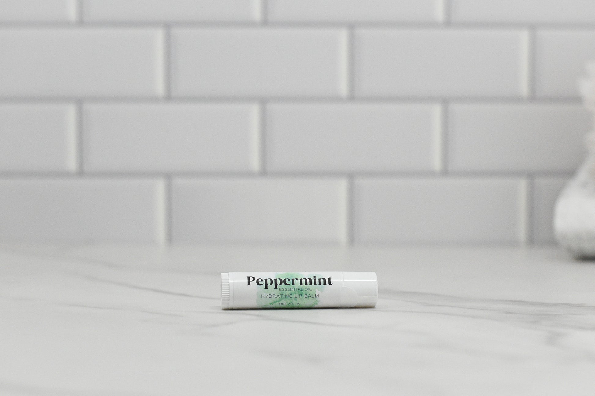 A tube of 570 Soap Co.'s Peppermint Essential Oil Lip Balm sits on a marble counter in front of a white subway tile backsplash, offering soothing hydration.