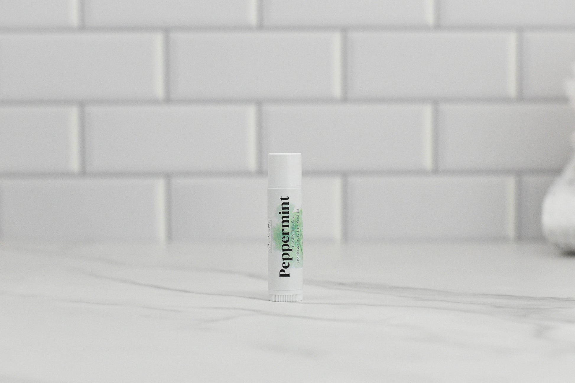 A tube of 570 Soap Co.'s Peppermint Essential Oil Lip Balm stands upright on a marble countertop, delivering rich hydration with every use. In the background, a white subway tile backsplash adds a classic touch, emphasizing the soothing presence of peppermint essential oil in each application.