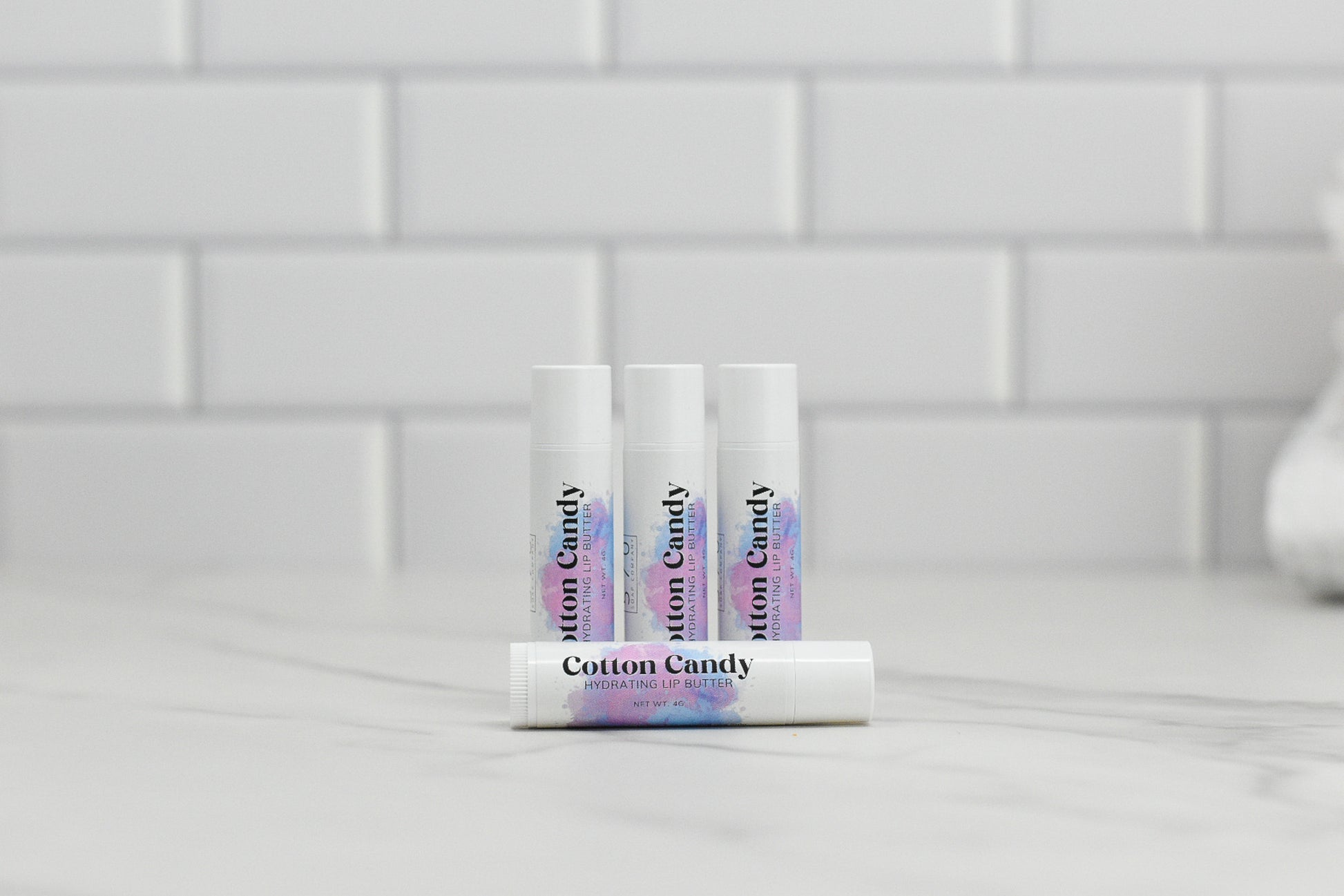 Four tubes of Cotton Candy Hydrating Lip Balm from 570 Soap Co. are arranged on a marble surface with a white tiled background. Made with natural ingredients, three tubes stand upright while one rests in front, displaying the product label.