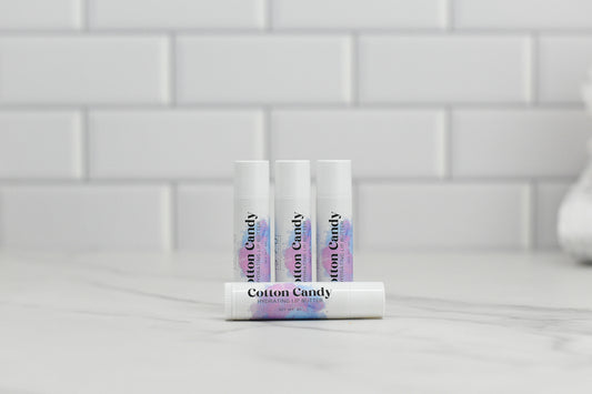 Four tubes of Cotton Candy Hydrating Lip Balm from 570 Soap Co. are arranged on a marble surface with a white tiled background. Made with natural ingredients, three tubes stand upright while one rests in front, displaying the product label.