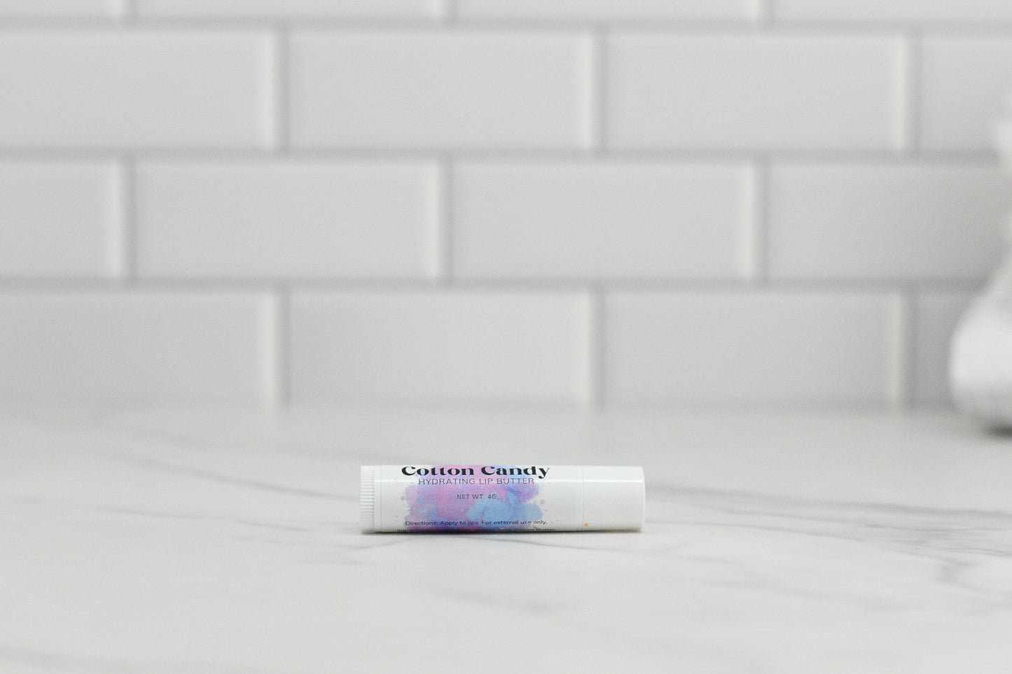 A small tube of 570 Soap Co.'s Cotton Candy Hydrating Lip Balm, crafted with natural ingredients, rests on a white marble countertop. The background showcases a wall adorned with white subway tiles.