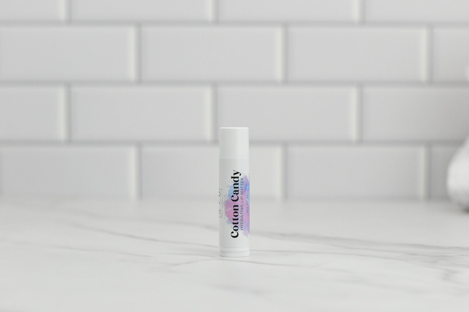 A tube of Cotton Candy Hydrating Lip Balm by 570 Soap Co., crafted with natural ingredients, stands on a marble countertop against a backdrop of white subway tiles.