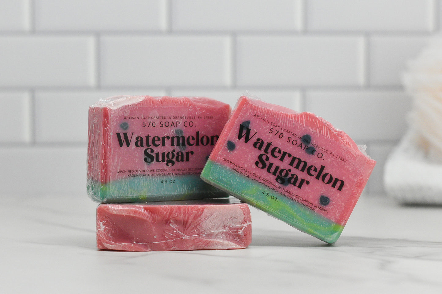 A trio of Sweet Watermelon Sugar Bar Soaps from 570 Soap Co. forms a fragrant ensemble as they rest atop a marble surface against a white tiled backdrop. These high-quality bars boast pink tops with green bases and seed-like accents, perfectly encapsulating the spirit of summer in soap form.
