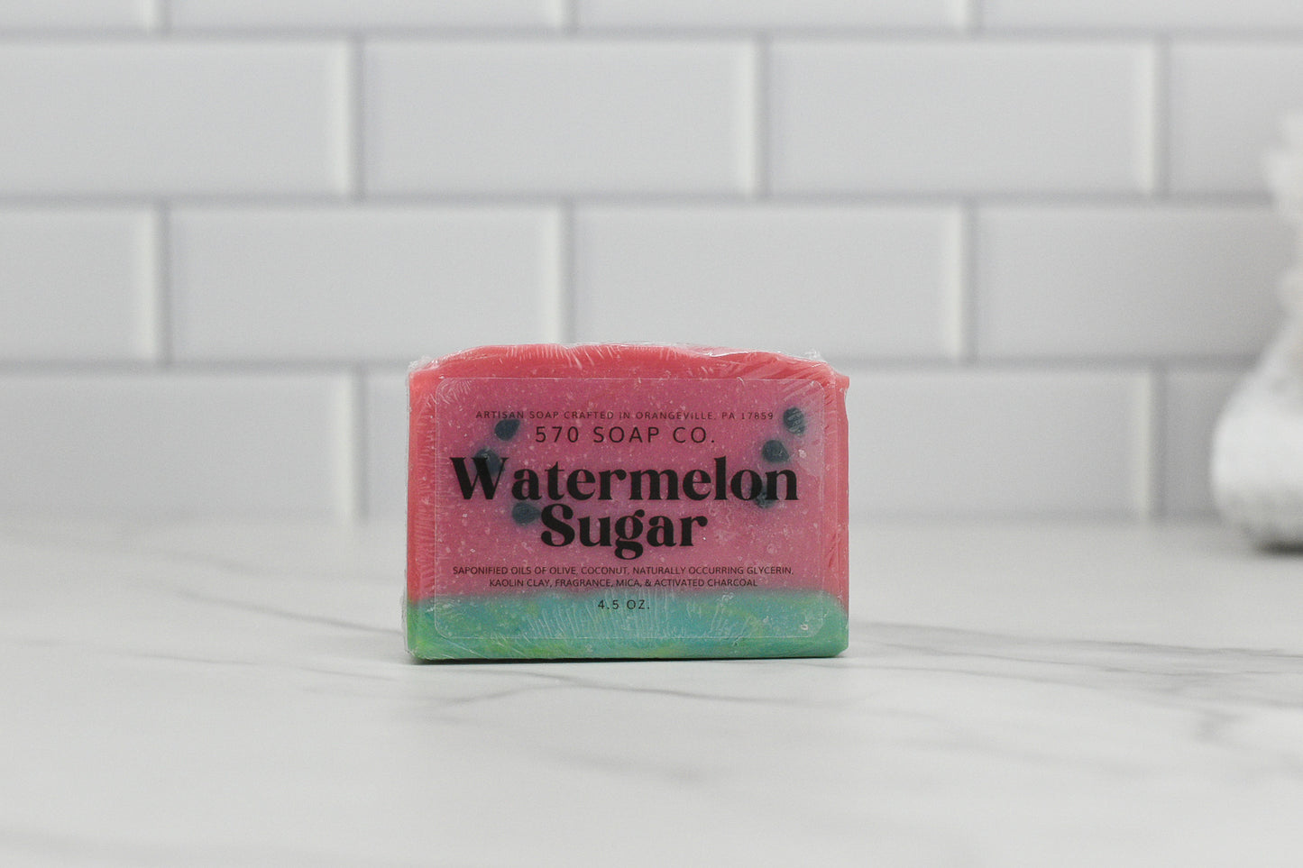 A bar of Sweet Watermelon Sugar soap from 570 Soap Co. sits elegantly on a marble surface against a backdrop of white tiled walls. This aromatic creation is designed in pink with a green base, beautifully resembling a slice of watermelon.