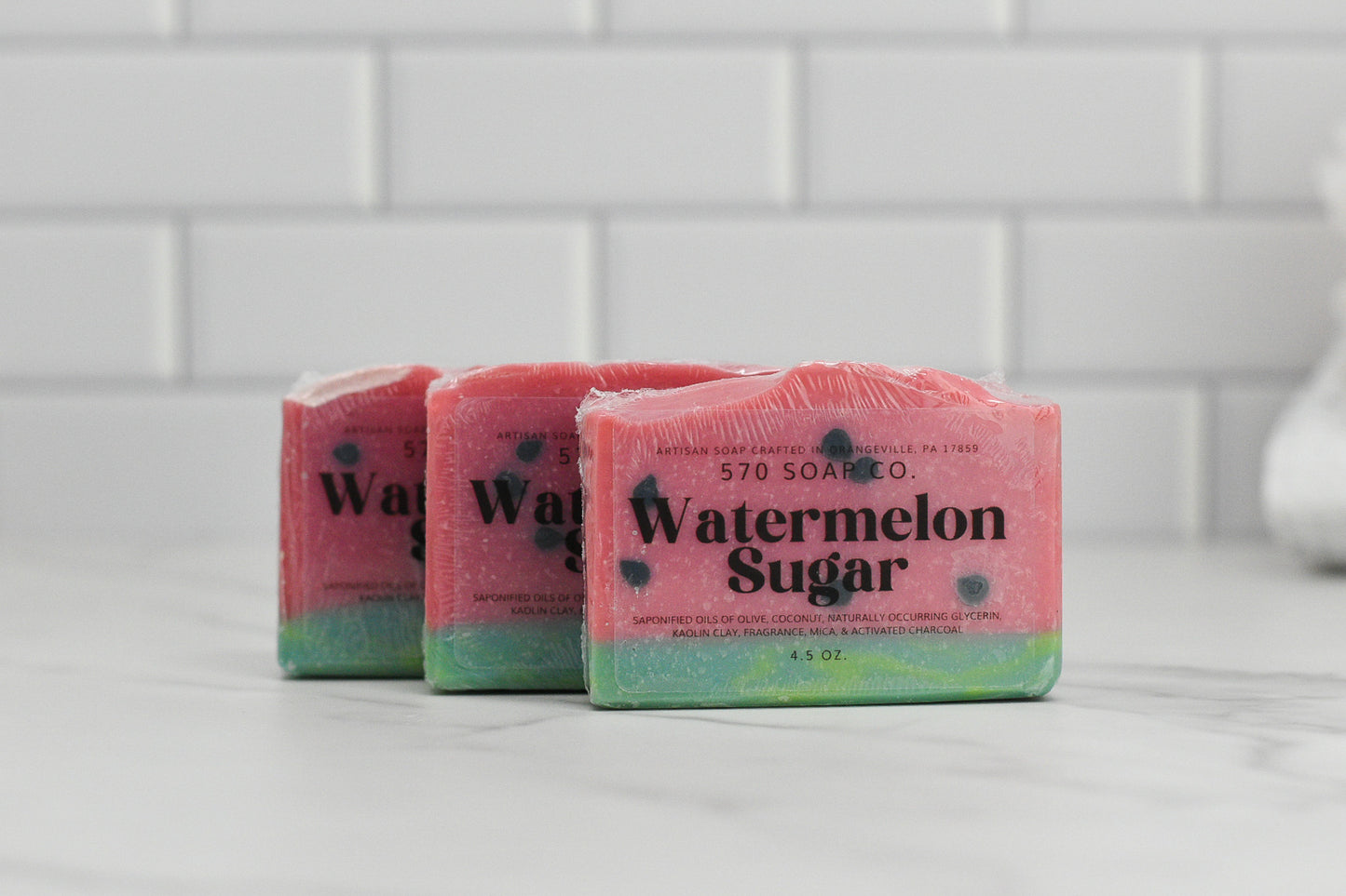 Three Sweet Watermelon Sugar Bar Soaps from 570 Soap Co. rest gracefully on a marble surface against a backdrop of white tiles. Each bar showcases a pink top adorned with black speckles and a green base, offering an aromatic experience that enchants the senses.