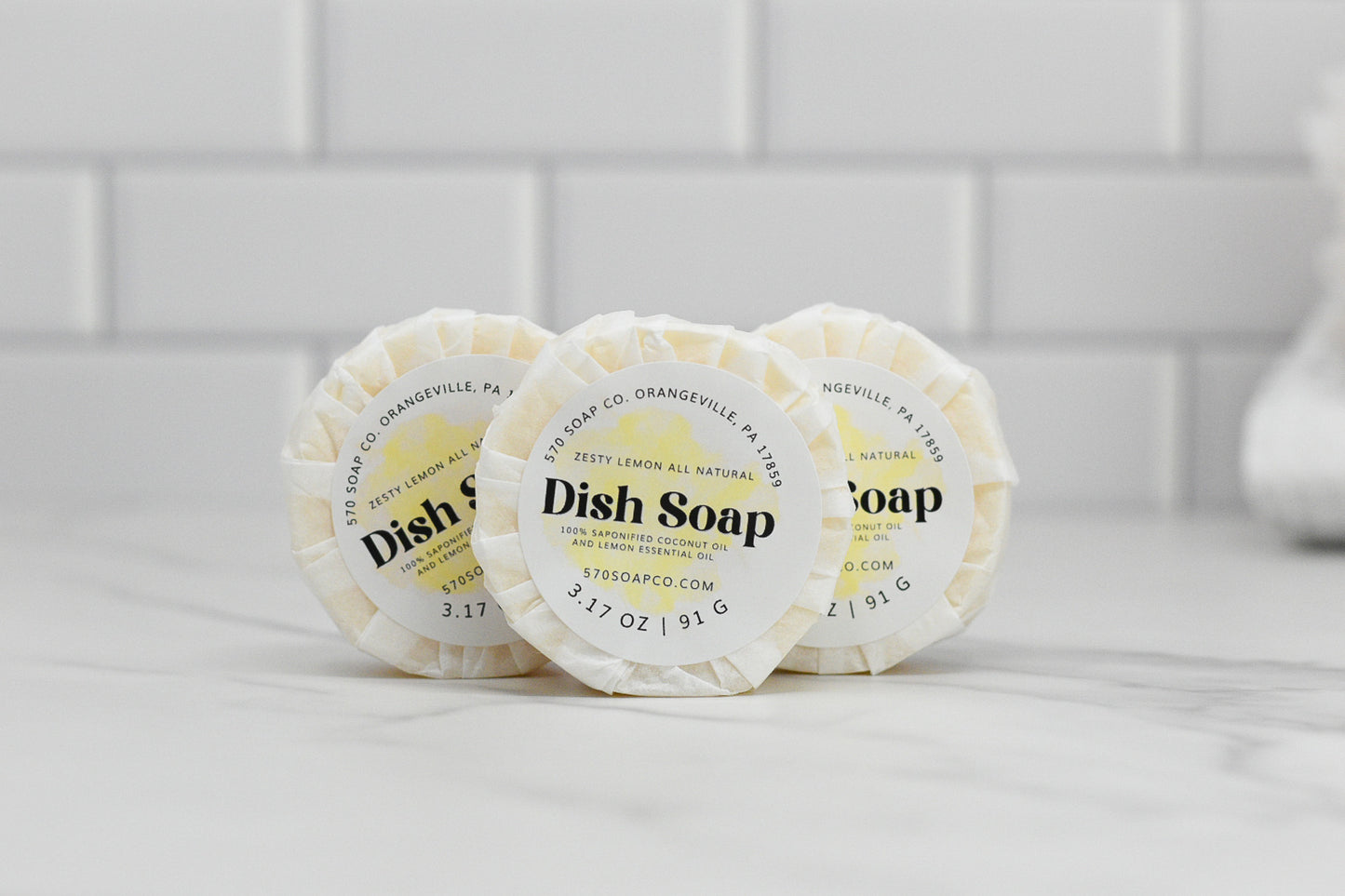 Three round bars of Zesty Lemon Dish Soap Bar - All Natural are neatly lined up on a white countertop against a tiled backsplash. Each eco-friendly kitchen essential from 570 Soap Co. features a label that reads "Dish Soap" and includes the ingredients as well as the brand's website. Delight in the fresh lemon scent while you tackle your dishes.