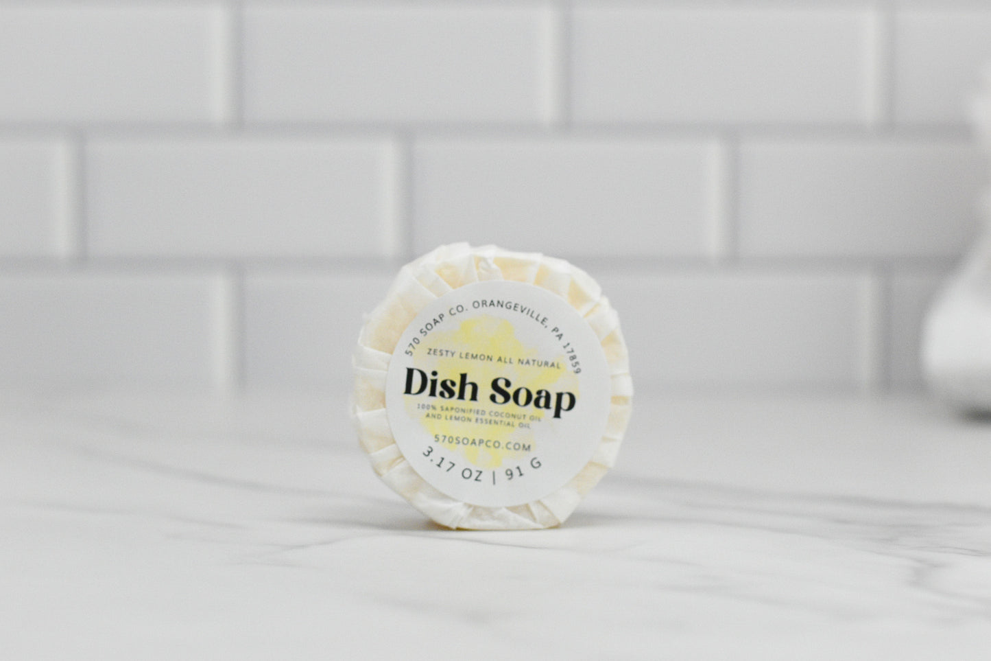 A round, wrapped Zesty Lemon Dish Soap Bar from 570 Soap Co. rests on a white countertop against a white tiled wall. With its refreshing lemon scent prominently advertised on the label, this all-natural solid dish soap weighs 3.17 oz (91g) and features a website below, making it an eco-friendly kitchen essential.