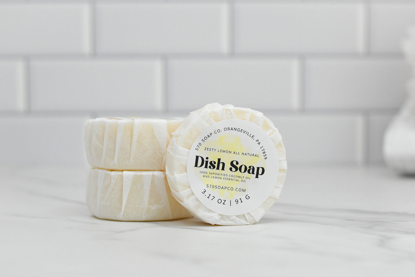 On a white marble countertop in front of a white tile backsplash, three bars of the all-natural Zesty Lemon Dish Soap from 570 Soap Co. are showcased. One bar is positioned on its side, displaying its label, while the other two are stacked flat, emphasizing this eco-friendly kitchen essential.