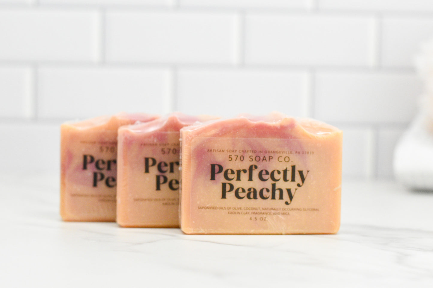 Three bars of Perfectly Peach Bar Soap: Exotic Fusion by 570 Soap Co. are elegantly displayed on a white marble surface. This soap showcases an intriguing blend of light and darker tones against a backdrop of white tiled walls, providing a skin-pampering experience that invigorates the senses.