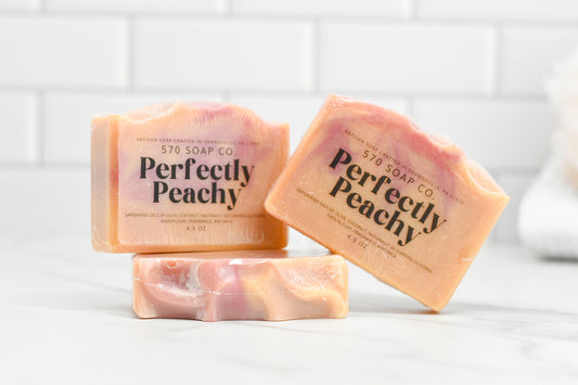 Three Perfectly Peach Bar Soaps: Exotic Fusion from 570 Soap Co. are displayed on a white countertop, showcasing their swirled peach and pink design. This soap boasts a skin-pampering formula, with the background featuring a white tiled wall and a blurred towel.