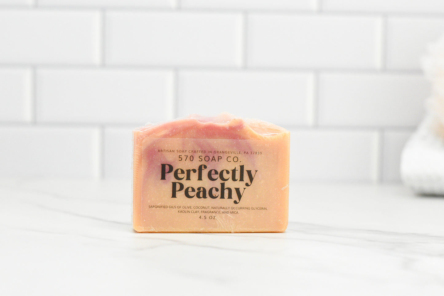 A bar of the luxurious Perfectly Peach Bar Soap: Exotic Fusion from 570 Soap Co. sits on a marble surface, highlighting its peach-toned beauty. The elegant white subway tile backdrop enhances the charm of this exquisite soap.