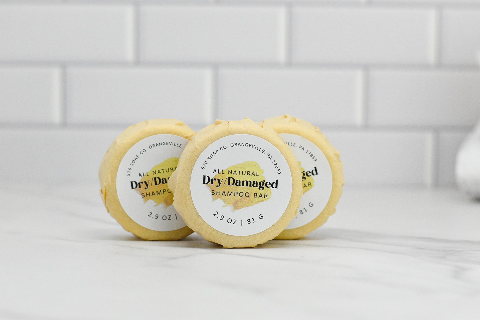 Three round Dry/Damaged Hair Shampoo Bars from 570 Soap Co. are displayed in a row on a white marble surface with a white tile background. Each all-natural bar, infused with essential oils, weighs 2.9 oz (81 g).
