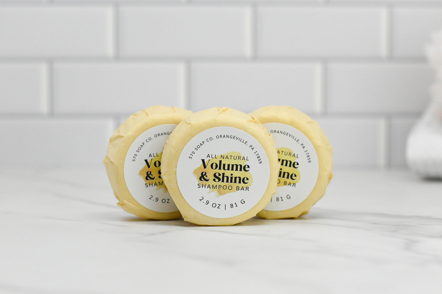 Three circular shampoo bars from 570 Soap Co., wrapped in yellow paper and labeled Volume & Shine Hair Shampoo Bar - All Natural, stand upright on a white countertop. Promising volume boost and radiant shine, they are set against a backdrop of a crisp white tiled wall.