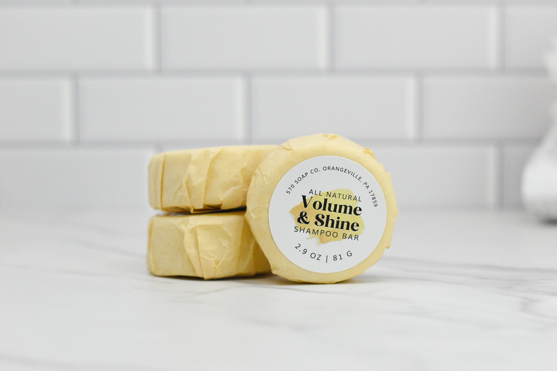 Three shampoo bars sit stacked on a white marble surface, each wrapped and displaying the label of the top bar: "Volume & Shine Hair Shampoo Bar - All Natural" by 570 Soap Co., weighing 2.9 oz / 81 g, with a promise of boosting volume and delivering radiant shine. Behind them is a backdrop of white tiled walls.