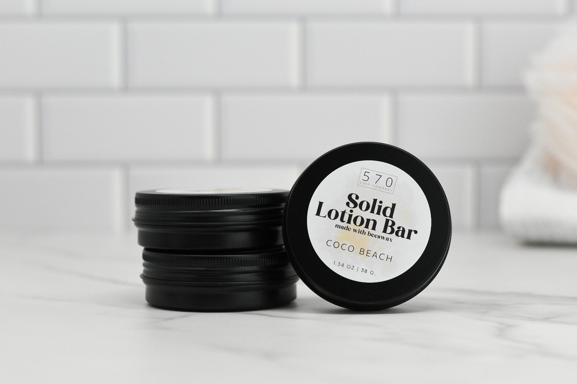 A stack of black containers, one marked with "Solid Lotion Bar - made with beeswax," captures attention. Highlighting its natural ingredients, the lid features the brand name 570 Soap Co. Ideal as a portable moisturizer, it rests against a white tile wall with a softly blurred object on the right.