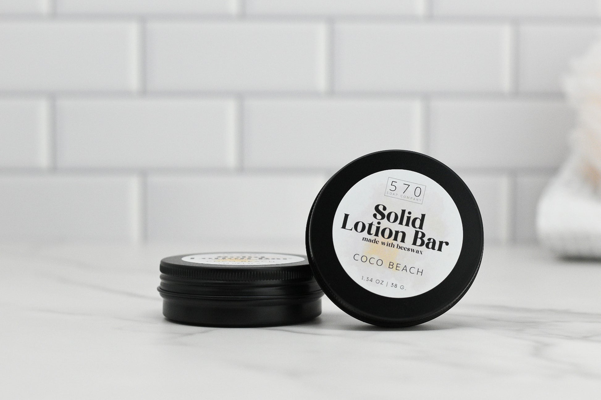 On a marble surface, two black tins from 570 Soap Co. are displayed; one is upright, revealing the label: Solid Lotion Bar - made with beeswax, Coco Beach. The other rests flat, highlighting this portable moisturizer crafted from natural ingredients against a backdrop of white tiled walls.