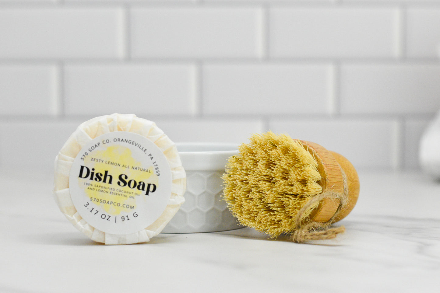 A block of Zesty Lemon Dish Soap Bar - All Natural, from 570 Soap Co., rests on a white marble counter next to a wooden dish brush with bristles. This eco-friendly kitchen essential is set against a backdrop of sleek white subway tiles.
