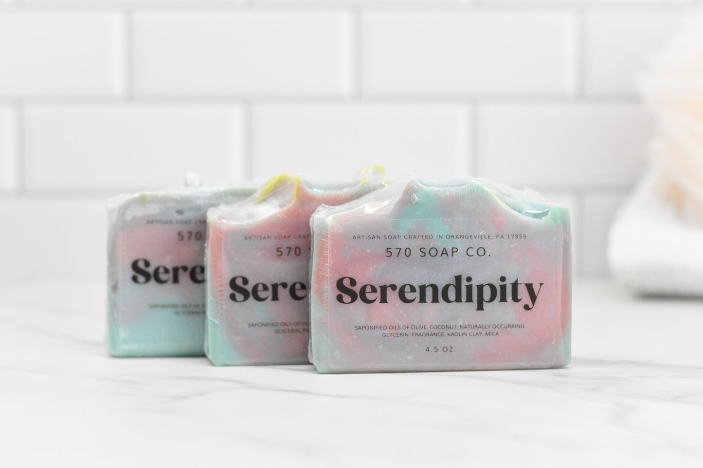 Three exquisite Serendipity Bar Soaps by 570 Soap Co. with their moisturizing formula are elegantly arranged on a white marble surface. The pastel shades of blue, pink, and green provide a vibrant contrast against the white subway tiles and the softly blurred backdrop of a towel.