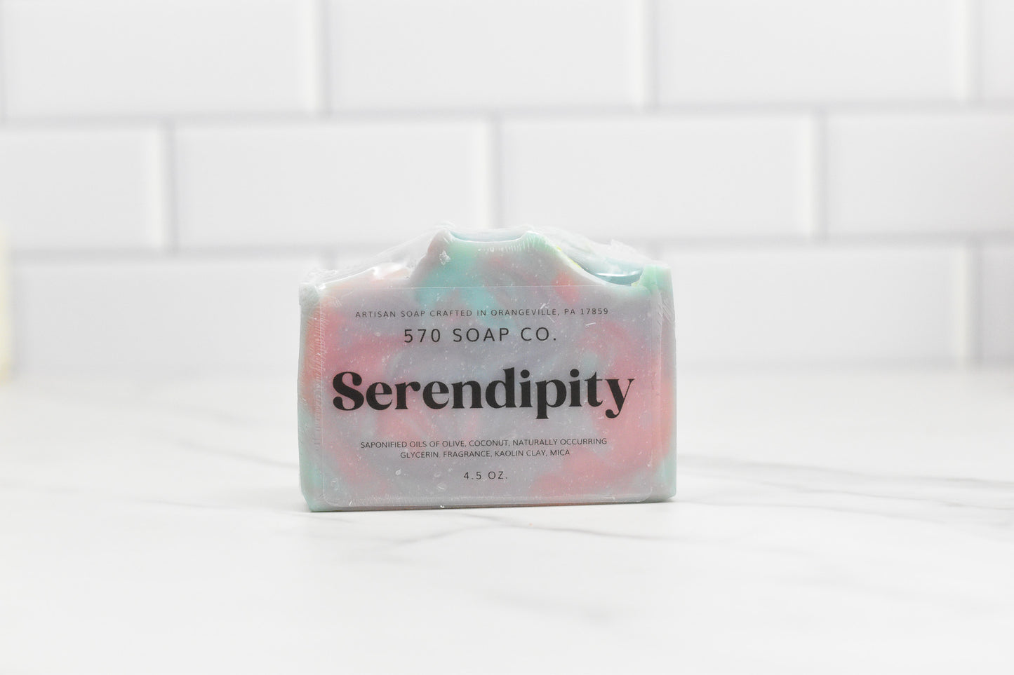 A luxurious Serendipity Bar Soap from 570 Soap Co. rests on a white countertop with a subway tile backdrop. Featuring a swirled pink and green design, this handcrafted soap boasts a moisturizing formula with gluten-free ingredients and locally sourced goat milk.